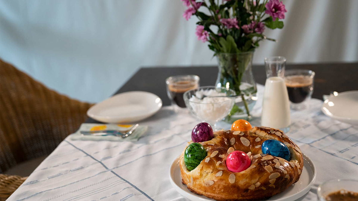 Ein festliches süßes Brot mit bunt gefärbten Eiern und Mandelscheiben steht auf einem mit Tellern, Kaffee, Gläsern und einer Vase mit lila Blumen gedeckten Tisch.