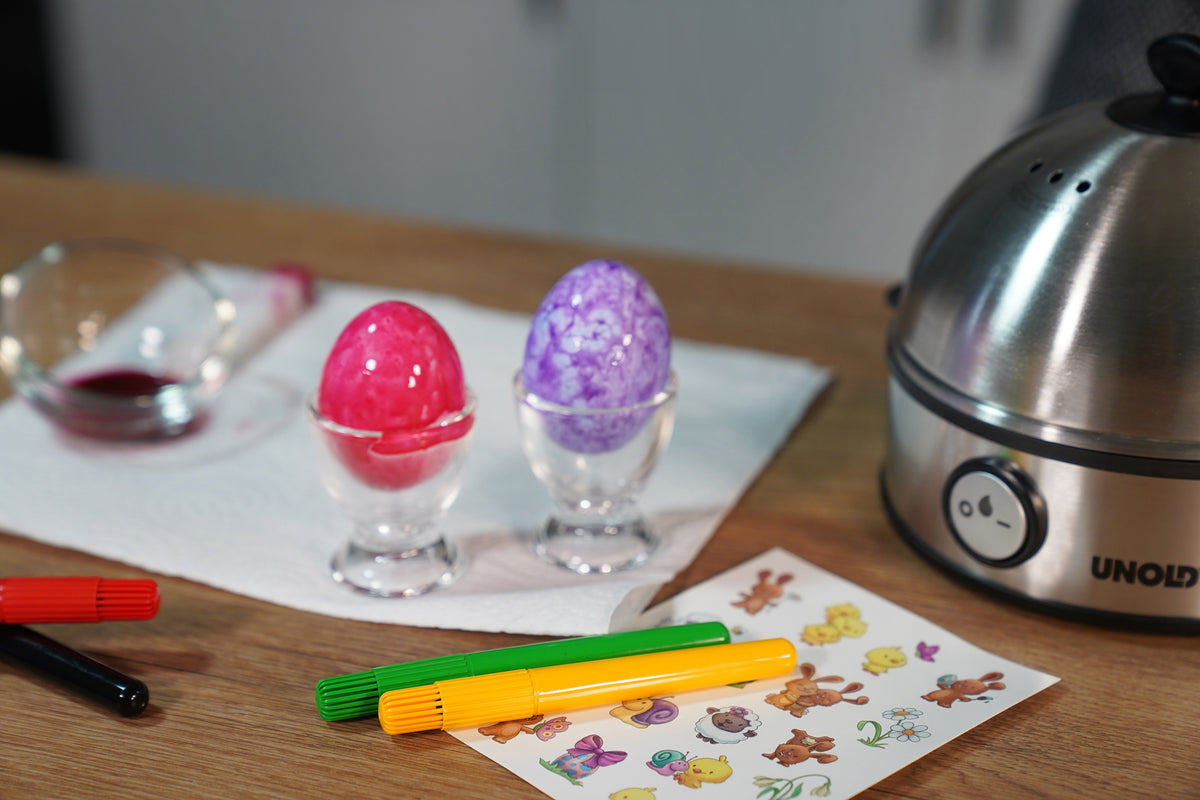Zwei bemalte Ostereier, ein rotes und ein violettes, liegen in Eierbechern aus Glas auf einem Papiertuch. Daneben liegen bunte Filzstifte, ein Bogen mit Tieraufklebern, eine Rührschüssel und ein Eierkocher auf einer Holzoberfläche.