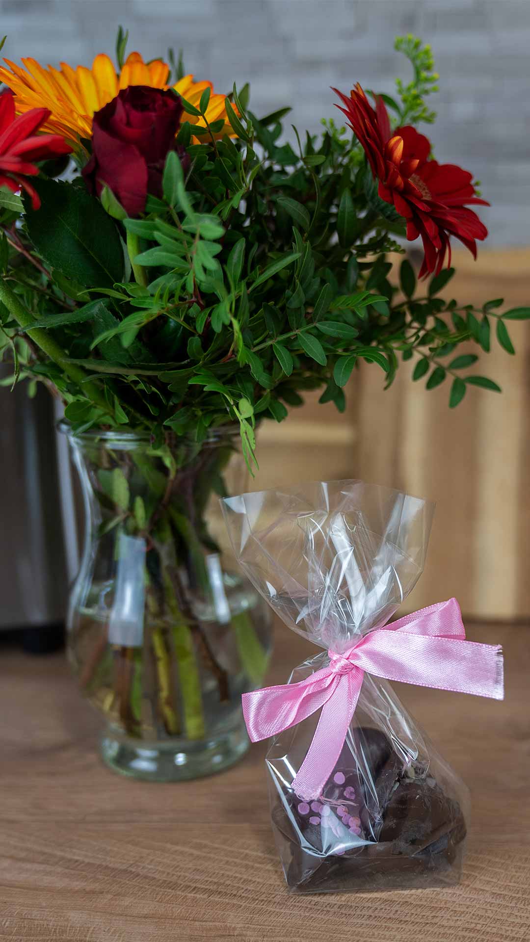Ein Strauß roter und orangefarbener Blumen in einer Glasvase steht auf einem Tisch neben einer durchsichtigen Plastiktüte mit Pralinen, die mit einer rosa Schleife zusammengebunden ist.
