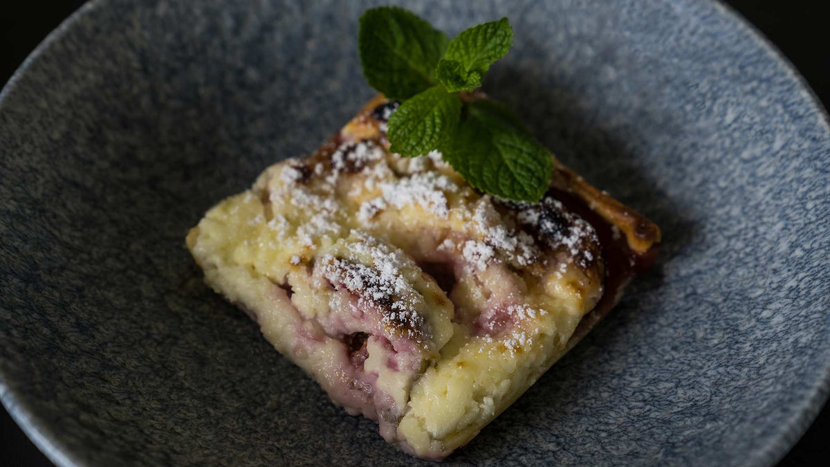Ein Stück Beeren-Crumble-Dessert, bestreut mit Puderzucker und garniert mit einem Zweig frischer Minze, serviert auf einem strukturierten grauen Teller.