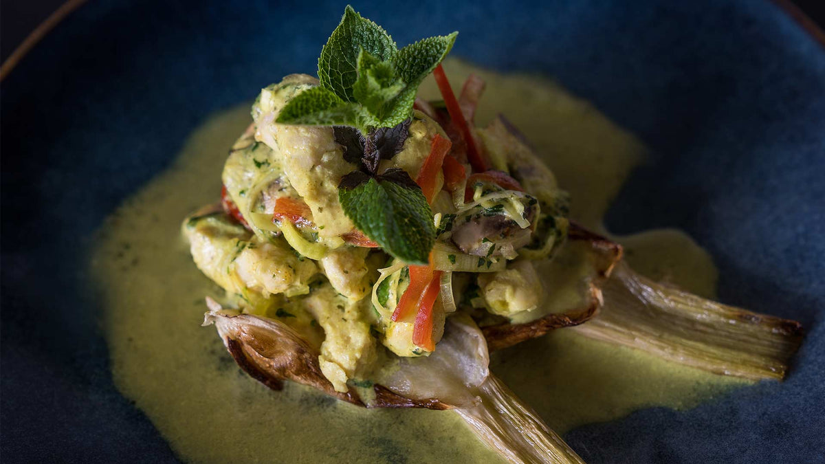 Ein Gourmetgericht mit Artischockenhälften, belegt mit einer cremigen Gemüse-Kräutermischung, garniert mit frischen Minzblättern, serviert auf einem dunkelblauen Teller mit grüner Sauce.