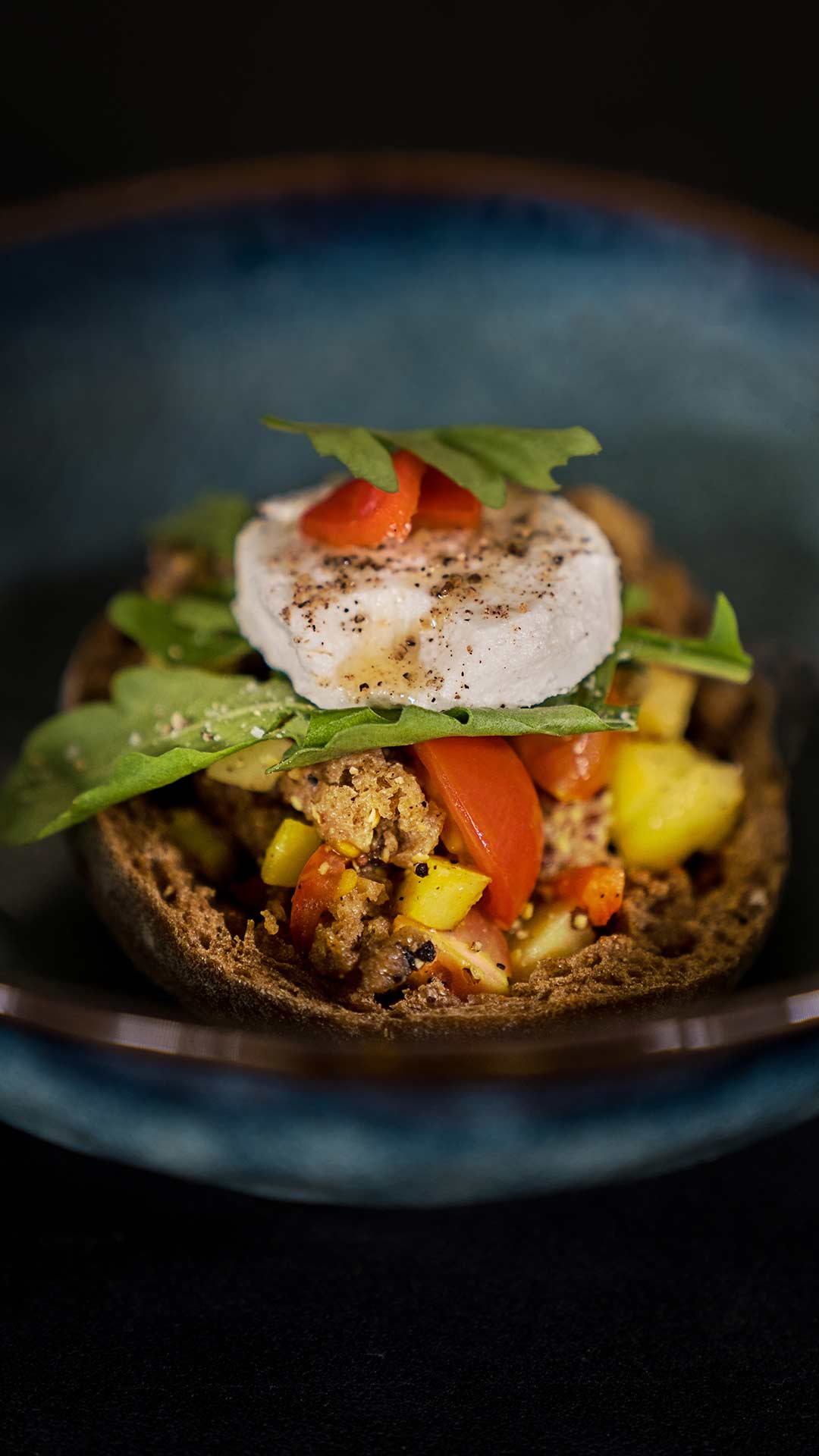 Eine Nahaufnahme eines Gourmetgerichts mit einer Scheibe Ziegenkäse, frischem Rucola, gewürfelten Paprika, Tomaten und gewürztem Hackfleisch auf einer Scheibe rustikalem Brot, serviert in einer tiefblauen Keramikschüssel.