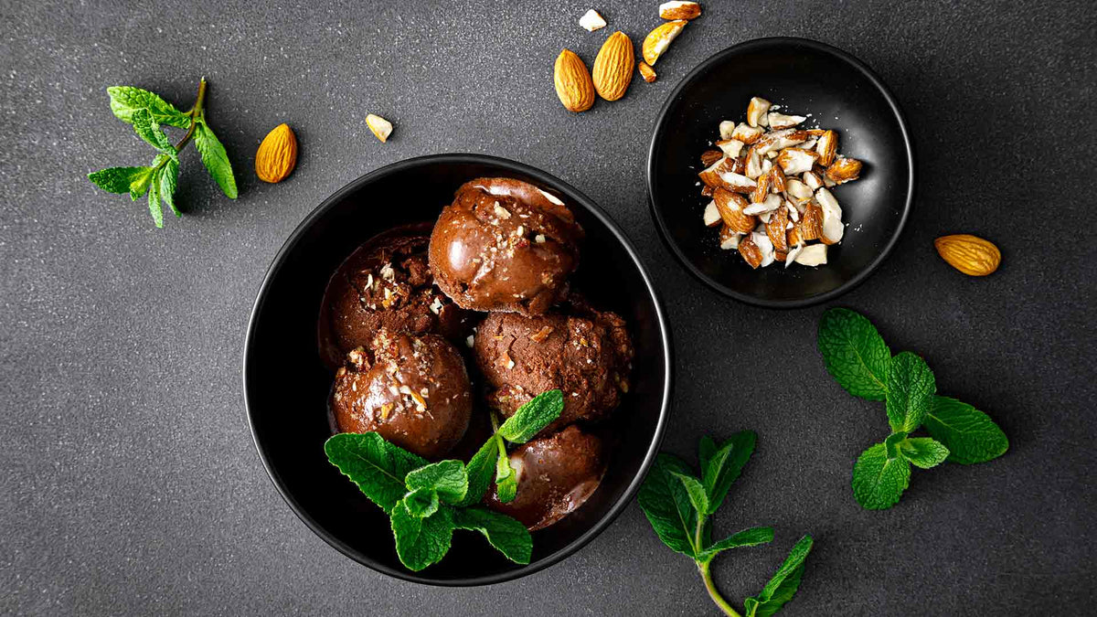 Eine schwarze Schüssel mit Kugeln Schokoladeneis, garniert mit Minzblättern und gehackten Mandeln, daneben eine kleine Schüssel mit gehackten Mandeln und verstreuten Minzzweigen und ganzen Mandeln auf einer dunklen Oberfläche.