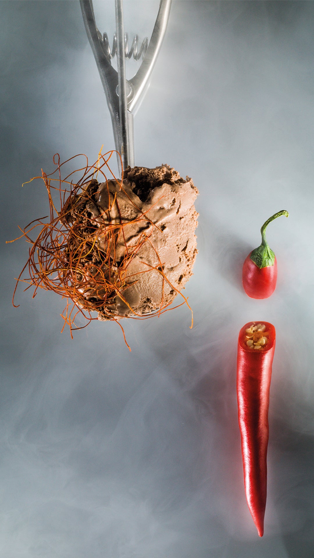 Eine Kugel Schokoladeneis mit Chilifäden in einem Metallportionierer steht neben einer ganzen roten Chilischote und einer geschnittenen Chilischote auf einem rauchgrauen Hintergrund.
