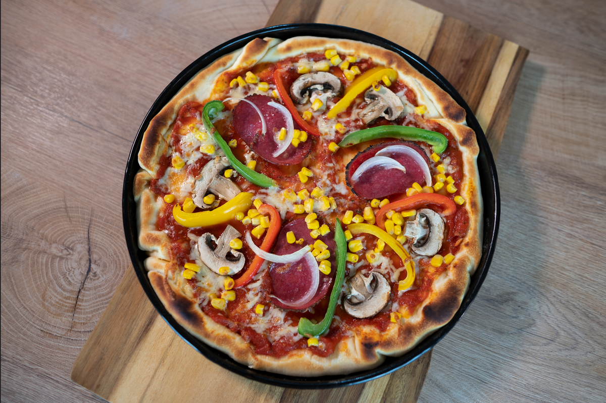Eine hausgemachte Pizza mit Peperoni, Pilzen, grünen und gelben Paprika, roten Zwiebeln und Mais auf einem schwarzen Blech auf einem Holzbrett.