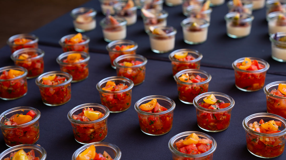 Auf einer schwarzen Tischdecke sind reihenweise kleine Gläser mit gewürfelten Tomaten und Kräutern, teilweise mit gelben Tomaten, angeordnet; im Hintergrund sind verschwommene Desserts in ähnlichen Gläsern zu sehen.