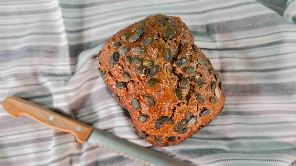 Ein mit Kürbiskernen bestreuter Laib Brot liegt auf einem gestreiften Tuch neben einem gezackten Messer mit Holzgriff.