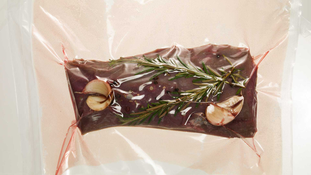 Ein rohes Steak wird mit Rosmarinzweigen, Knoblauchzehen und gemahlenem Pfeffer in einem Vakuumbeutel versiegelt und ist bereit zum Sous-Vide-Garen.