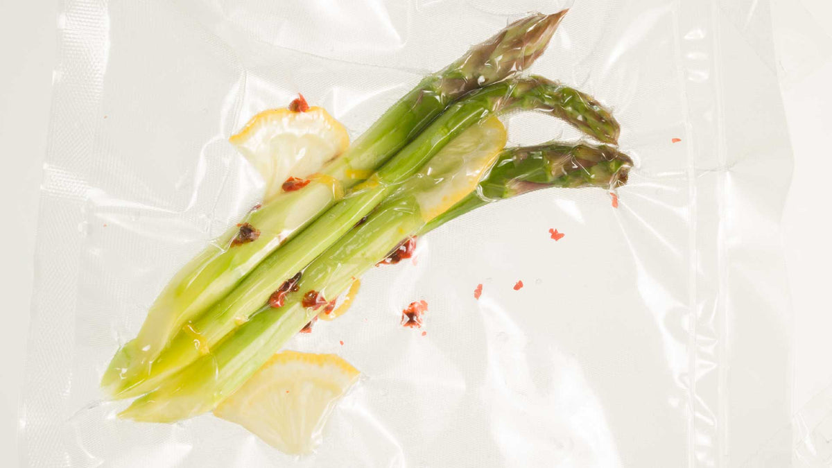 Drei Spargelstangen mit Zitronenscheiben und roten Pfefferflocken sind in einem durchsichtigen Sous-Vide-Vakuumbeutel vor einem schlichten weißen Hintergrund versiegelt.