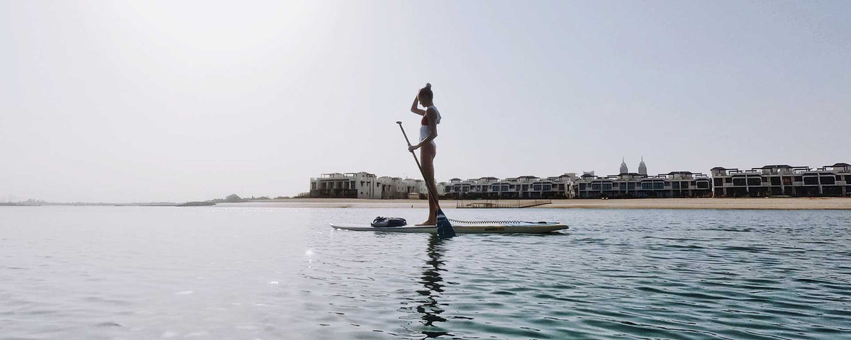 Eine Frau im Badeanzug steht auf einem Paddleboard in ruhigem Wasser in Ufernähe, im Hintergrund sind moderne Gebäude und ein klarer Himmel zu sehen.
