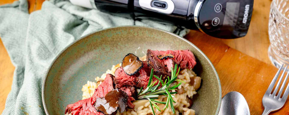 Eine Schüssel cremiges Risotto, garniert mit Scheiben von blutigem Steak, schwarzem Trüffel und einem Zweig Rosmarin, neben einem Küchengerät und einem Glas auf einem Holztisch.