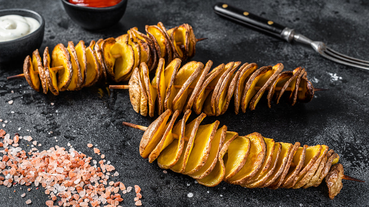 Auf einer dunklen Oberfläche werden zwei Spieße mit spiralförmig geschnittenen Bratkartoffeln präsentiert. Daneben stehen Schüsseln mit weißen und roten Dip-Saucen, eine Gabel und eine Prise rosa Himalaya-Salz.