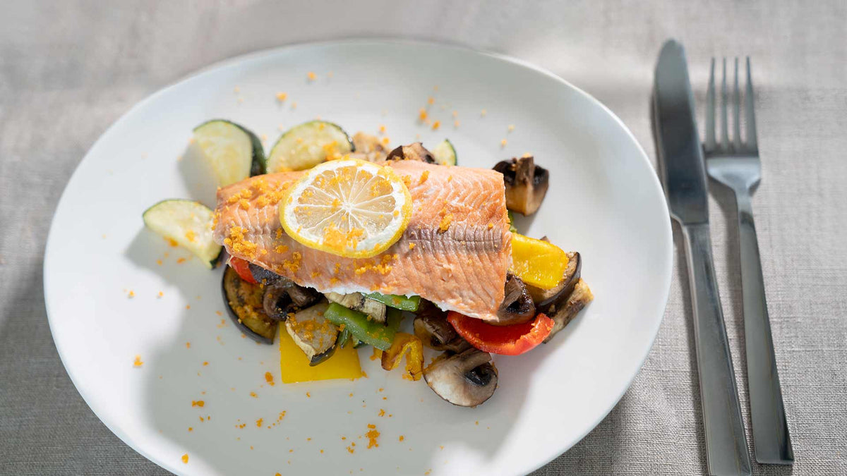 Ein Teller mit gebackenem Lachs, garniert mit einer Zitronenscheibe, serviert auf einem Bett aus geröstetem Gemüse, darunter Zucchini, Paprika und Pilze. Messer und Gabel liegen auf einer grauen Stoffserviette neben dem Teller.