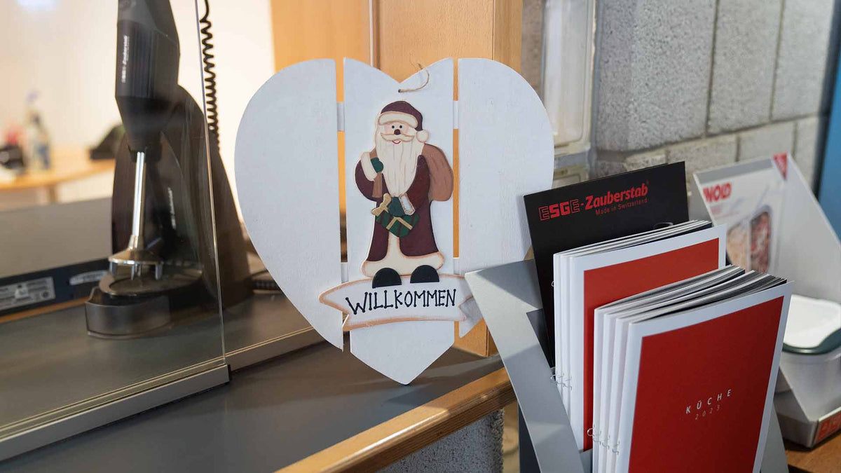 Ein herzförmiges Holzschild mit einer Weihnachtsmannfigur und dem Wort „Willkommen“ hängt auf einem Tresen neben einem Prospektständer mit rot-weißen Heften.