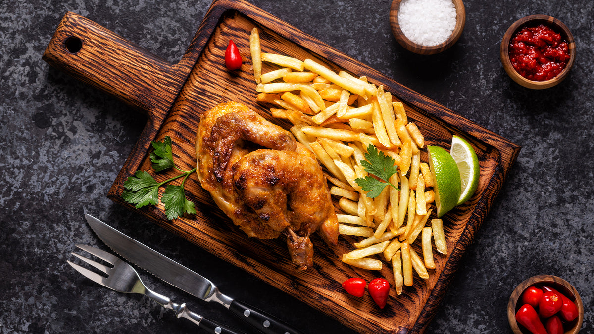 Gegrilltes Hähnchen mit Pommes Frites, Limettenspalten, Petersilie und kleinen roten Paprikaschoten auf einem Holzbrett. Gabel, Messer, Salz und rote Sauce liegen daneben auf einer dunklen Steinoberfläche.