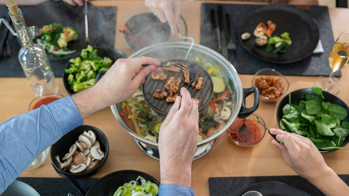Die Leute versammelten sich um einen Tisch und kochten Fleisch und Gemüse auf einem Tischgrill. Um den heißen Topf herum waren Teller mit frischem Gemüse, Pilzen und Dip-Saucen angeordnet.