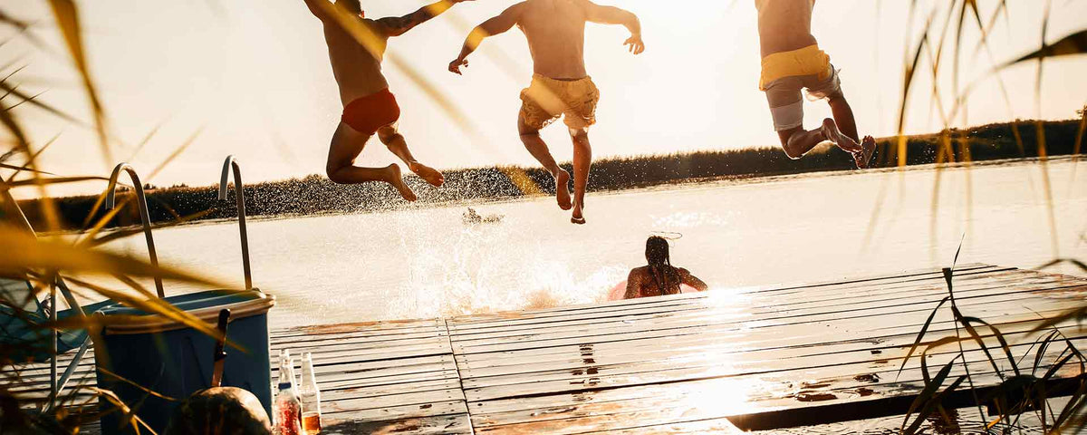 Drei Personen in Badebekleidung springen von einem Holzsteg in einen sonnenbeschienenen See, während eine weitere Person bereits im Wasser liegt. Die Szene ist hell und sommerlich, mit plätscherndem Wasser und warmem Sonnenlicht.