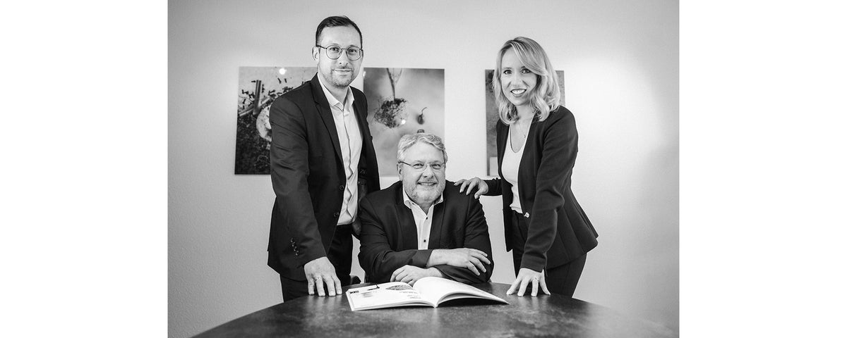 Drei Personen in Business-Kleidung posieren gemeinsam in einem Büro. Zwei stehen lächelnd rechts und links eines sitzenden Mannes. Vor ihnen liegt eine aufgeschlagene Zeitschrift auf dem Schreibtisch. An der Wand hinter ihnen hängen zwei abstrakte Kunstwerke.