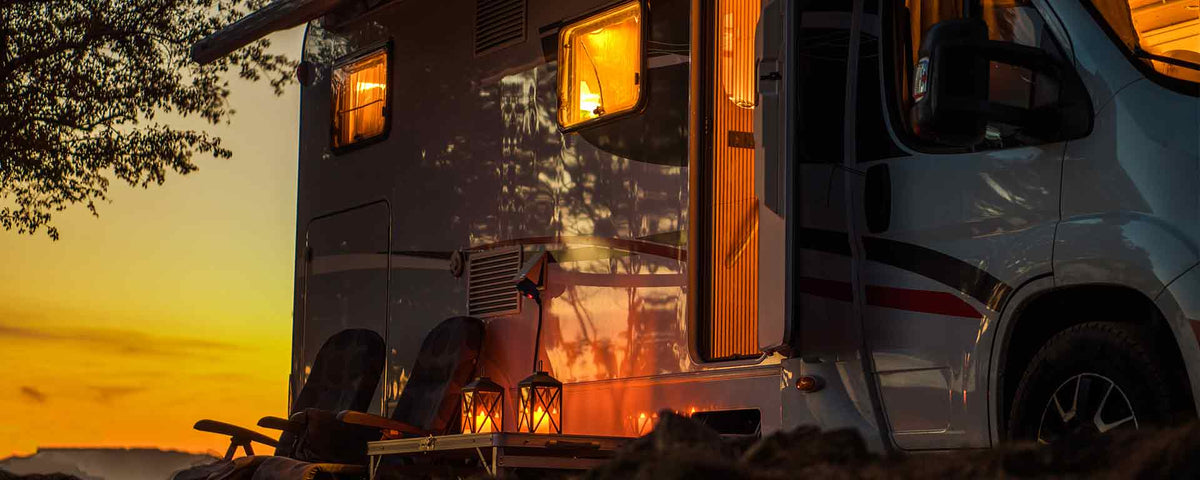 Ein gemütlicher Wohnwagen steht bei Sonnenuntergang draußen, von innen leuchtet warmes Licht, draußen sind zwei Stühle und ein kleiner Tisch mit Laternen aufgestellt und die Silhouette eines Baums zeichnet sich gegen den bunten Himmel ab.