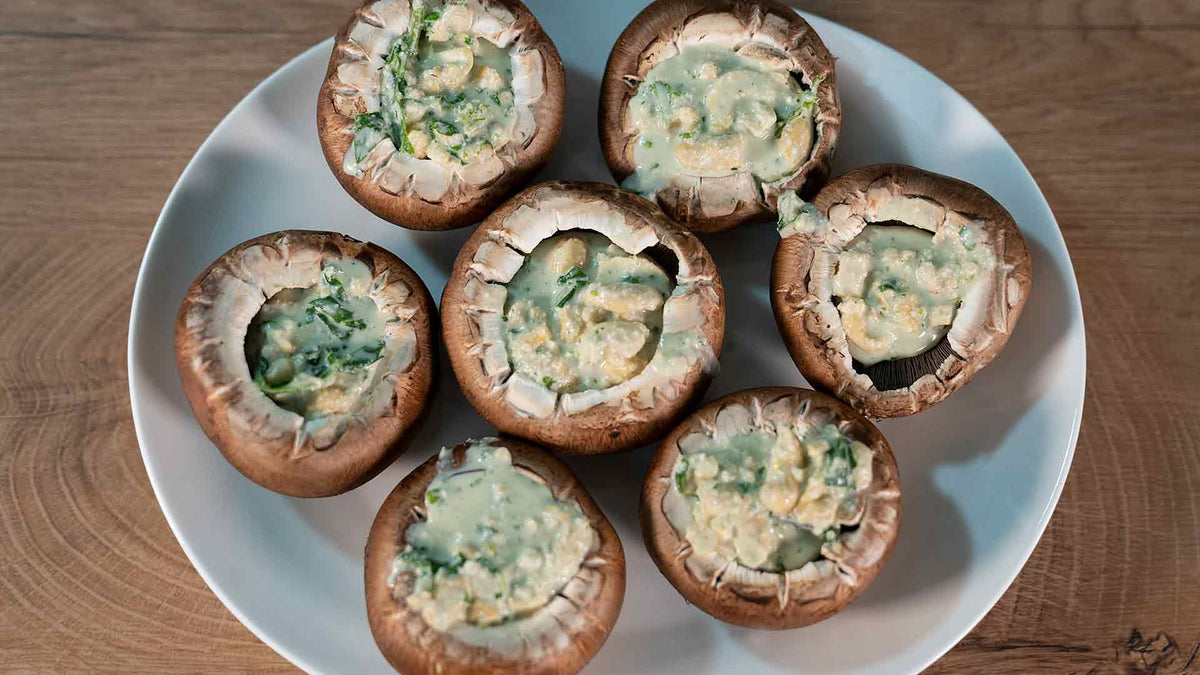 Auf einem weißen Teller liegen sieben große, mit einer cremigen Käse-Kräuter-Mischung gefüllte Champignons auf einer Holzoberfläche.
