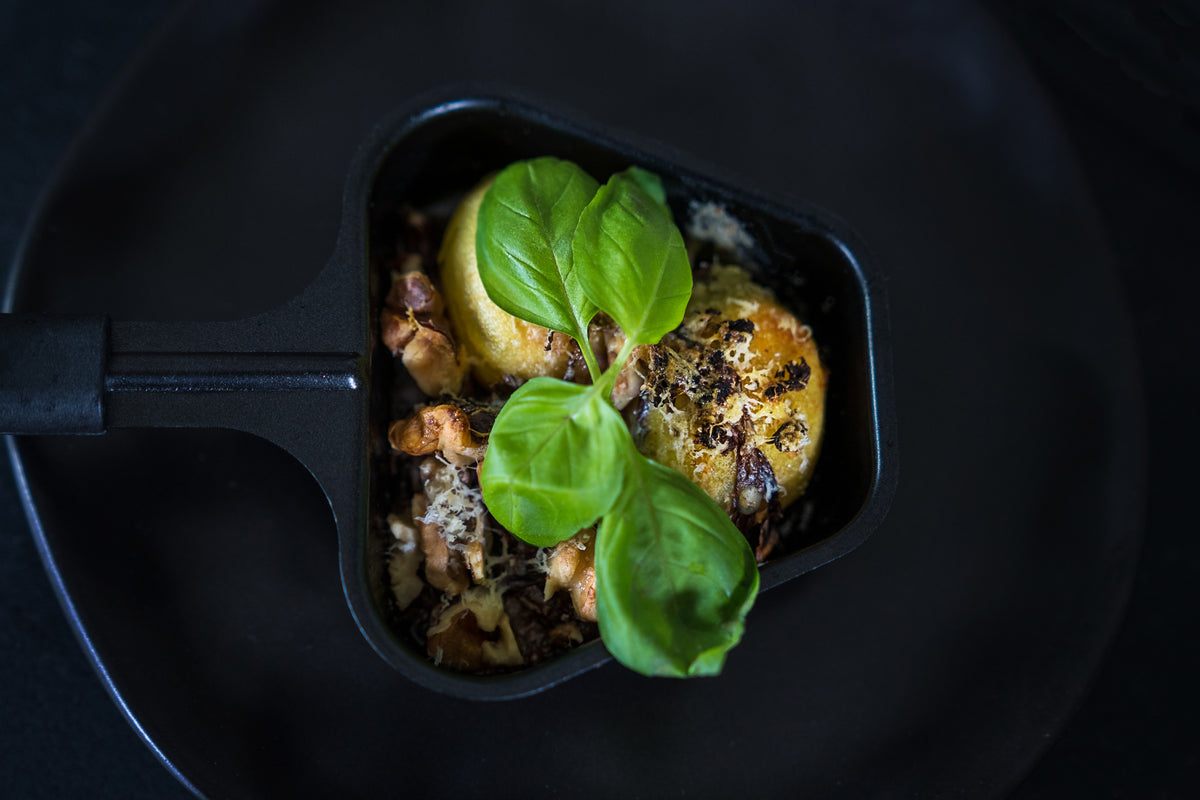 Das Unold Raclette Rezeptbuch Deluxe zeigt eine kleine schwarze Pfanne mit Ofenkartoffeln, frischem Basilikum, gehackten Nüssen und Käse, alles elegant serviert auf einem dunklen Teller.