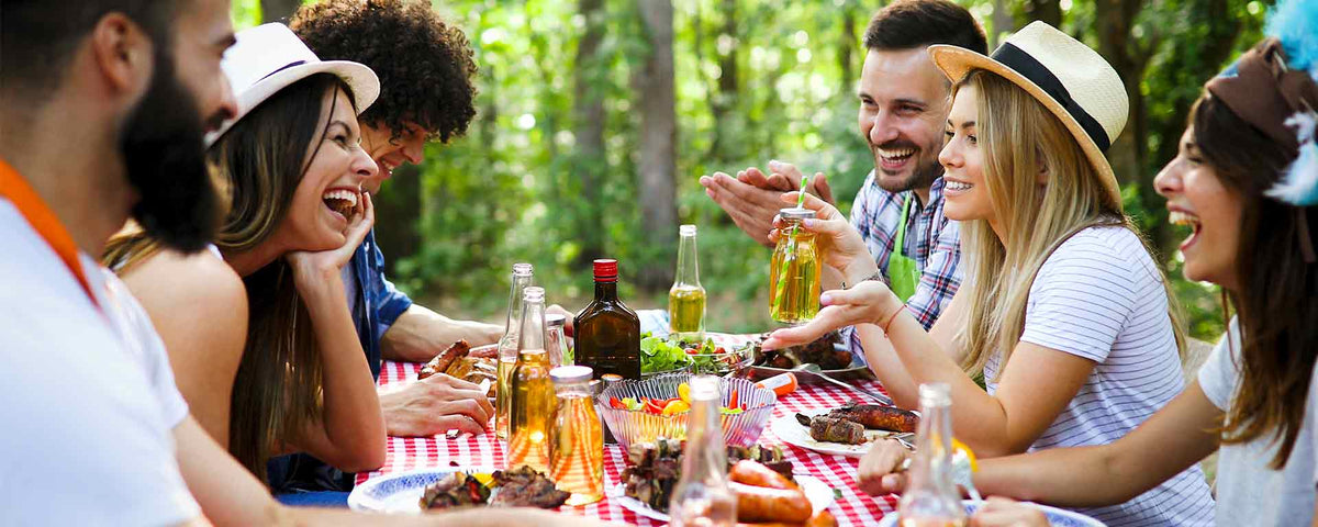 Eine Gruppe von Freunden sitzt an einem sonnigen Tag im Freien um einen Picknicktisch, lacht und genießt gemeinsam Essen und Getränke. Der Tisch ist mit einem rot karierten Tuch bedeckt und von Bäumen umgeben.