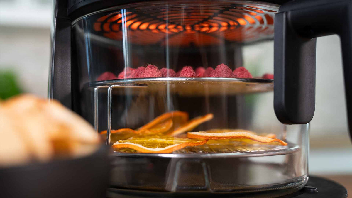 Eine Nahaufnahme eines Dörrgeräts im Einsatz. Zitrusfruchtscheiben und kleine, runde, rote Stücke werden auf separaten Tabletts unter einem glühenden Heizelement getrocknet.