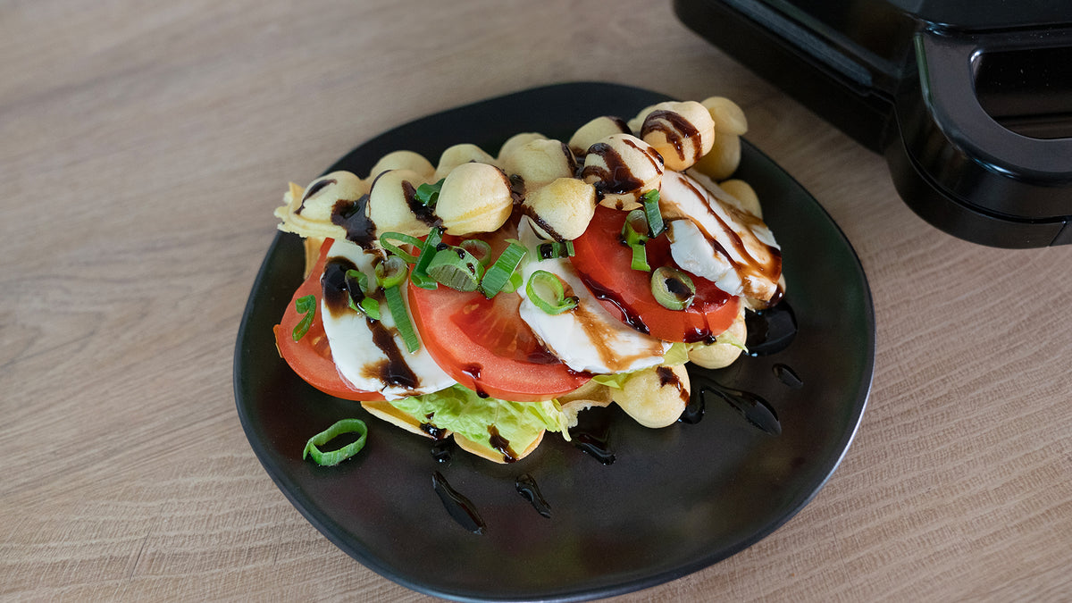 Eine Bubble-Waffel mit Salat, Tomatenscheiben, Mozzarella, Frühlingszwiebeln und Balsamico-Glasur, serviert auf einem schwarzen Teller auf einem Holztisch.