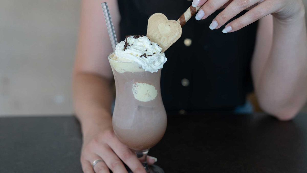 Eine Person hält ein Glas Schokoladenmilchshake mit Schlagsahne, Schokoladenraspeln, einem herzförmigen Keks und einem Waffelstäbchen in der Hand. Die Person hat manikürte Nägel und trägt ein dunkles ärmelloses Top.
