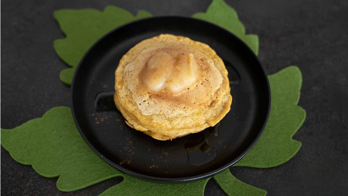 Ein Stapel Pfannkuchen mit einem Stück Butter und Sirup liegt auf einem schwarzen Teller mit einem grünen, blattförmigen Tischset darunter vor einem dunklen Hintergrund.