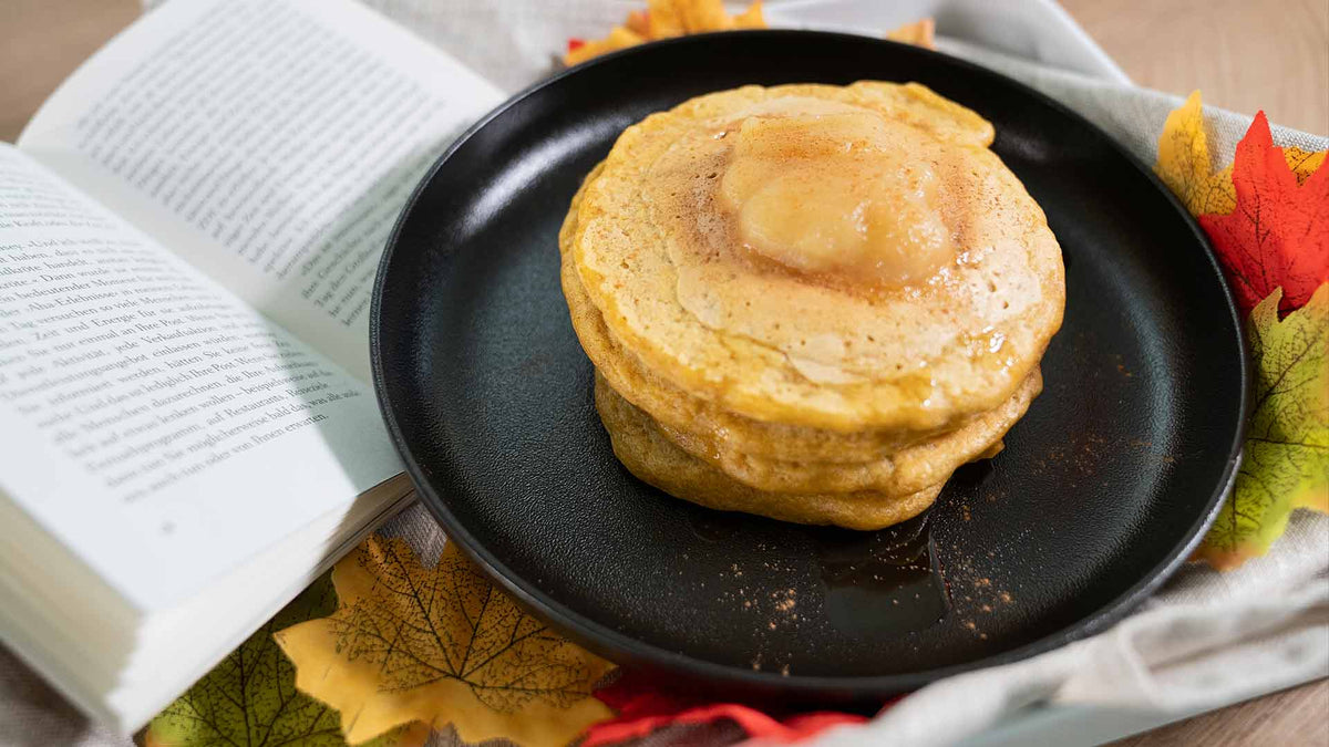 Ein Stapel Pfannkuchen mit Butter darauf liegt auf einem schwarzen Teller, umgeben von Herbstblättern und einem offenen Buch, was eine gemütliche Herbstatmosphäre schafft.