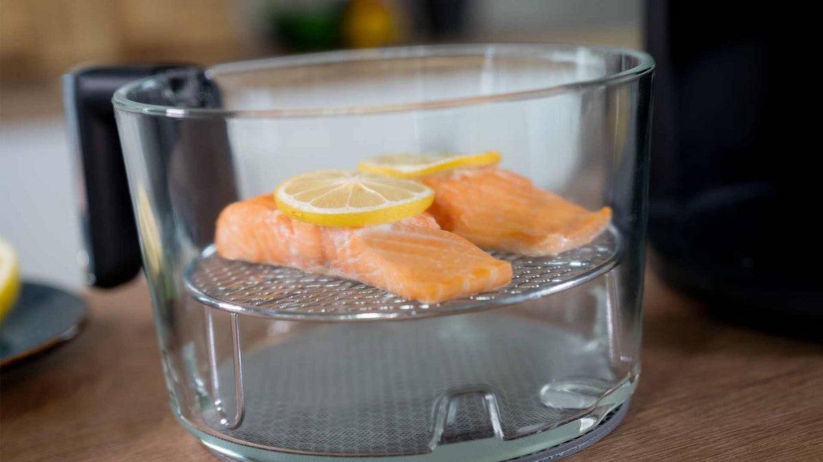 Zwei rohe Lachsfilets mit Zitronenscheiben obenauf liegen auf einem Metallrost in einem Glaskorb der Heißluftfritteuse und sind bereit zum Kochen.