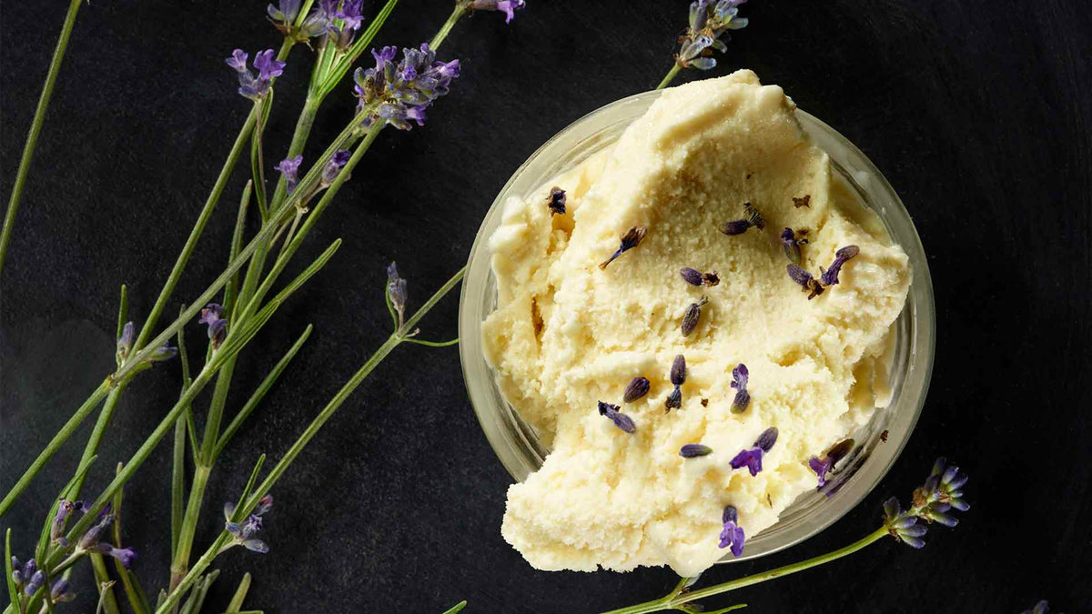 Eine Schale mit cremigem Vanilleeis, garniert mit violetten Lavendelknospen, auf einer dunklen Oberfläche neben Zweigen mit frischen Lavendelblüten platziert.