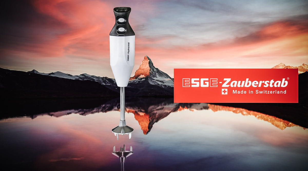 Ein weißer ESGE-Zauberstab-Stabmixer steht vor einer malerischen Berglandschaft mit einem spiegelnden See und rosa Himmel, neben einem roten ESGE-Zauberstab-Logo „Made in Switzerland“.