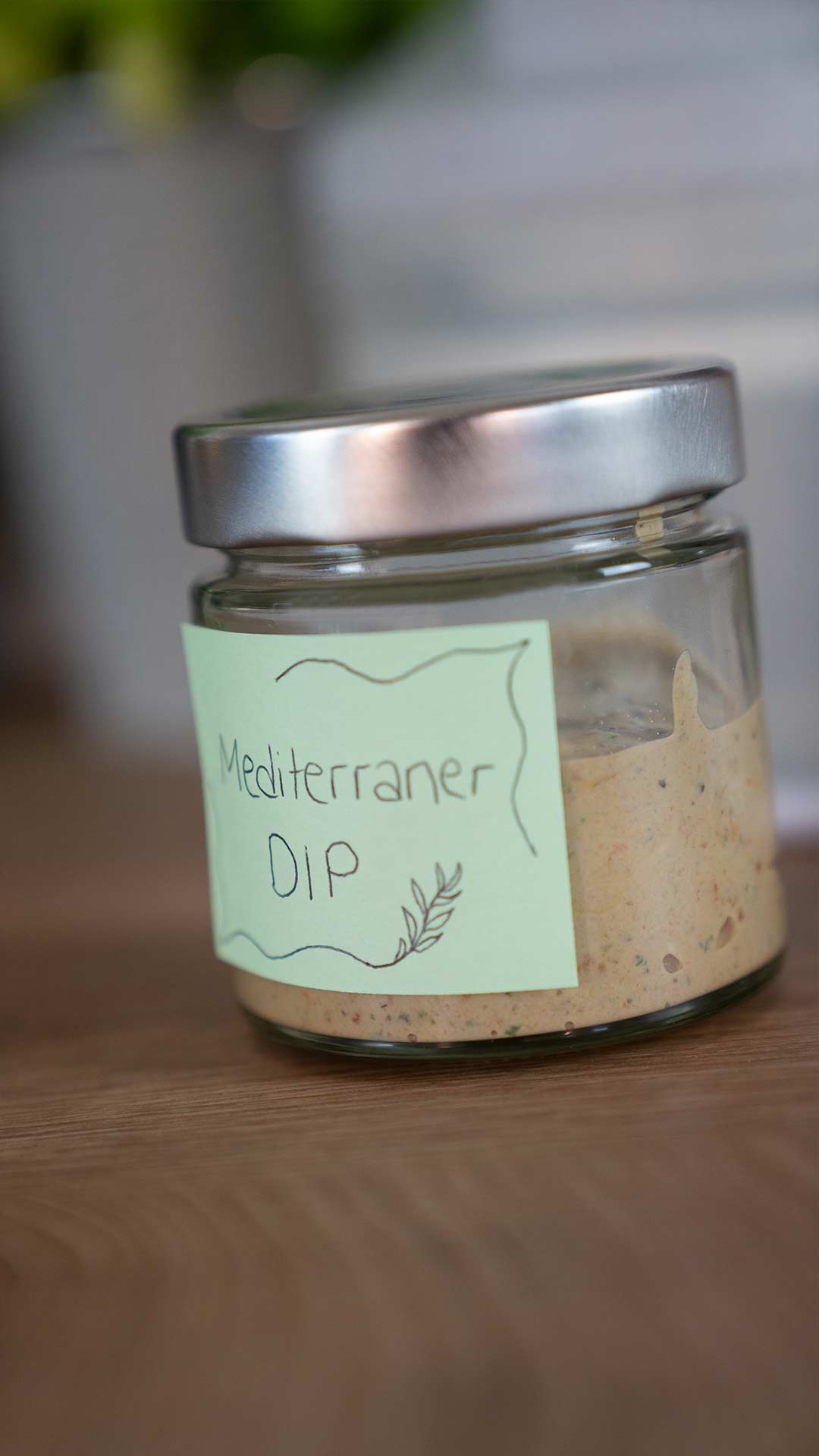 Ein kleines Glasgefäß mit silbernem Deckel steht auf einer Holzfläche. Das handgeschriebene Etikett auf hellgrünem Papier trägt die Aufschrift „Mediterraner Dip“ und ist mit einer schlichten Blattillustration verziert. Das Glas ist teilweise mit einem beigen Dip gefüllt.