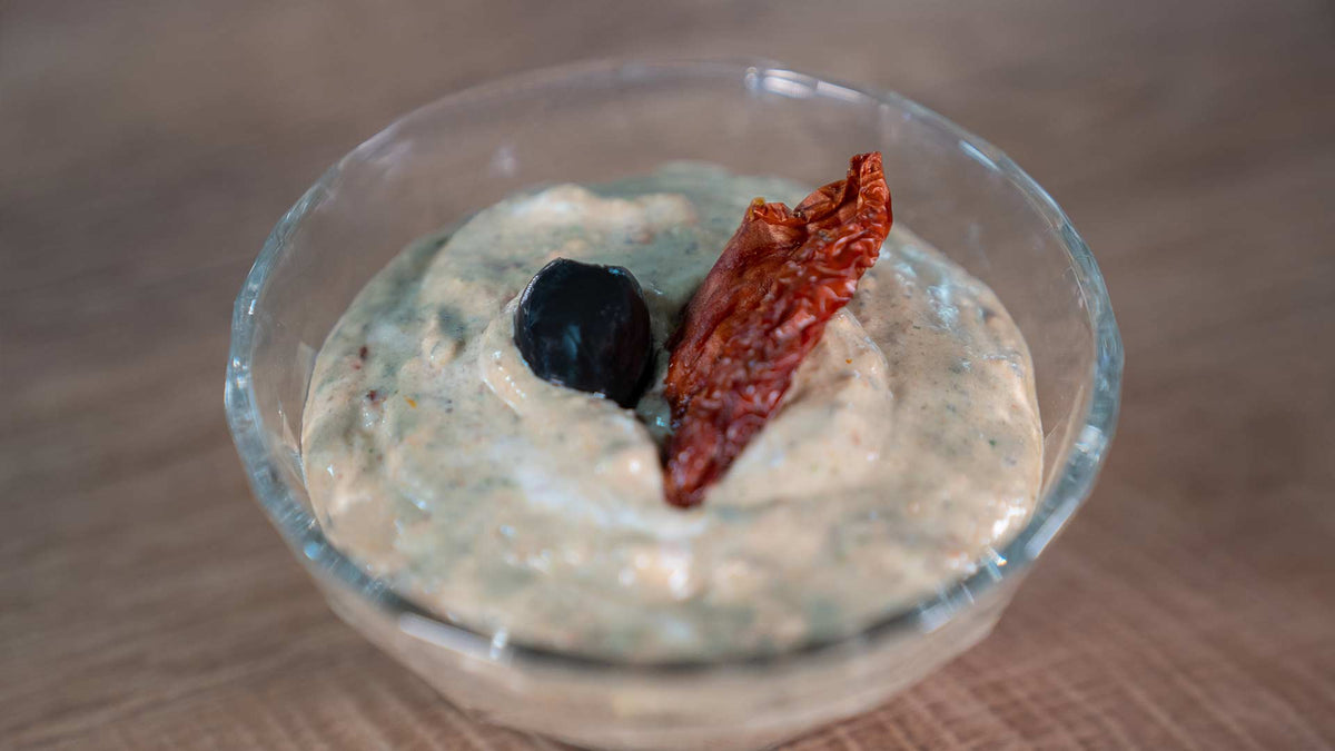 Eine Glasschale gefüllt mit cremigem Dip, garniert mit einer schwarzen Olive und einer Scheibe sonnengetrockneter Tomate, platziert auf einer Holzoberfläche.