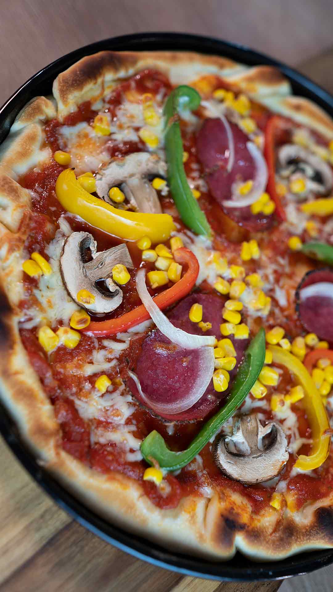 Eine Nahaufnahme einer Pizza mit gelbem Mais, geschnittenen Champignons, grünen und gelben Paprika, roten Zwiebeln, Peperoni und geschmolzenem Käse auf einem knusprigen Boden.