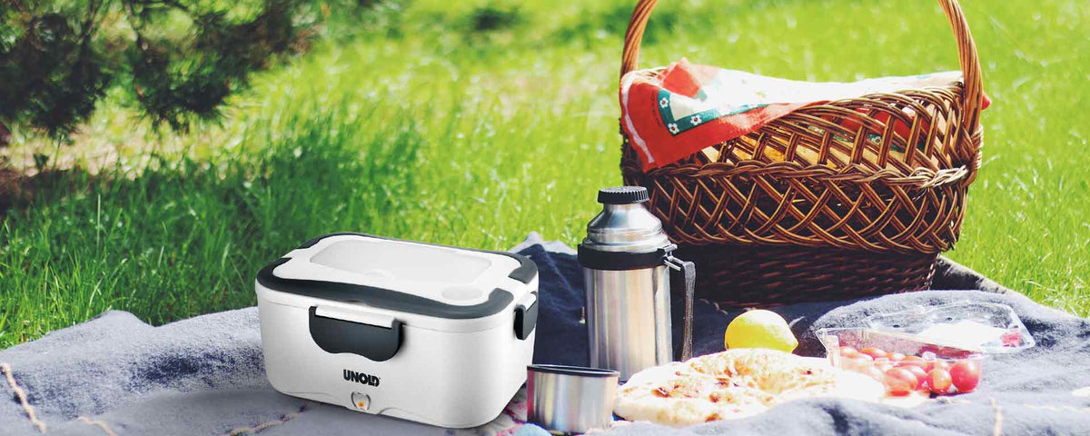 Ein Picknick-Setup auf Gras mit einer Decke, einem Weidenkorb, einer Thermoskanne, einer Tasse, Obst, Trauben, Pizza und einem tragbaren weißen UNOLD-Speisenwärmer im Vordergrund.