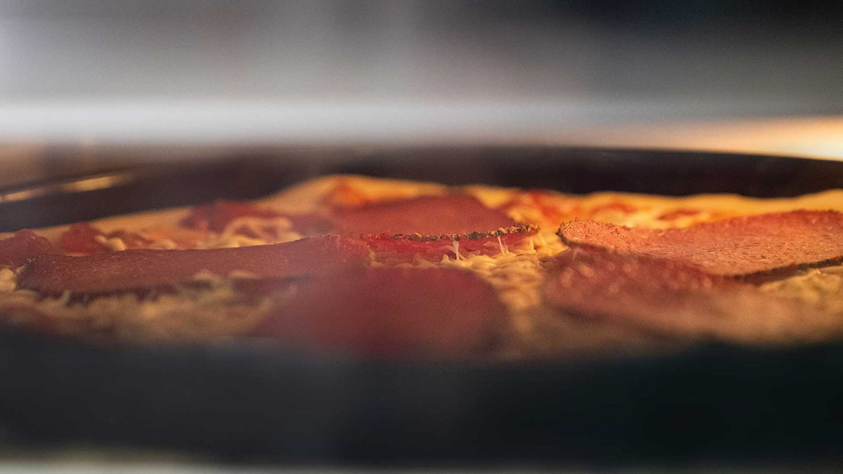 Nahaufnahme einer Pizza im Ofen. Auf dem Rand sind Peperonischeiben und geschmolzener Käse zu sehen. Das Bild ist leicht unscharf, was die Hitze und Atmosphäre im Ofen hervorhebt.