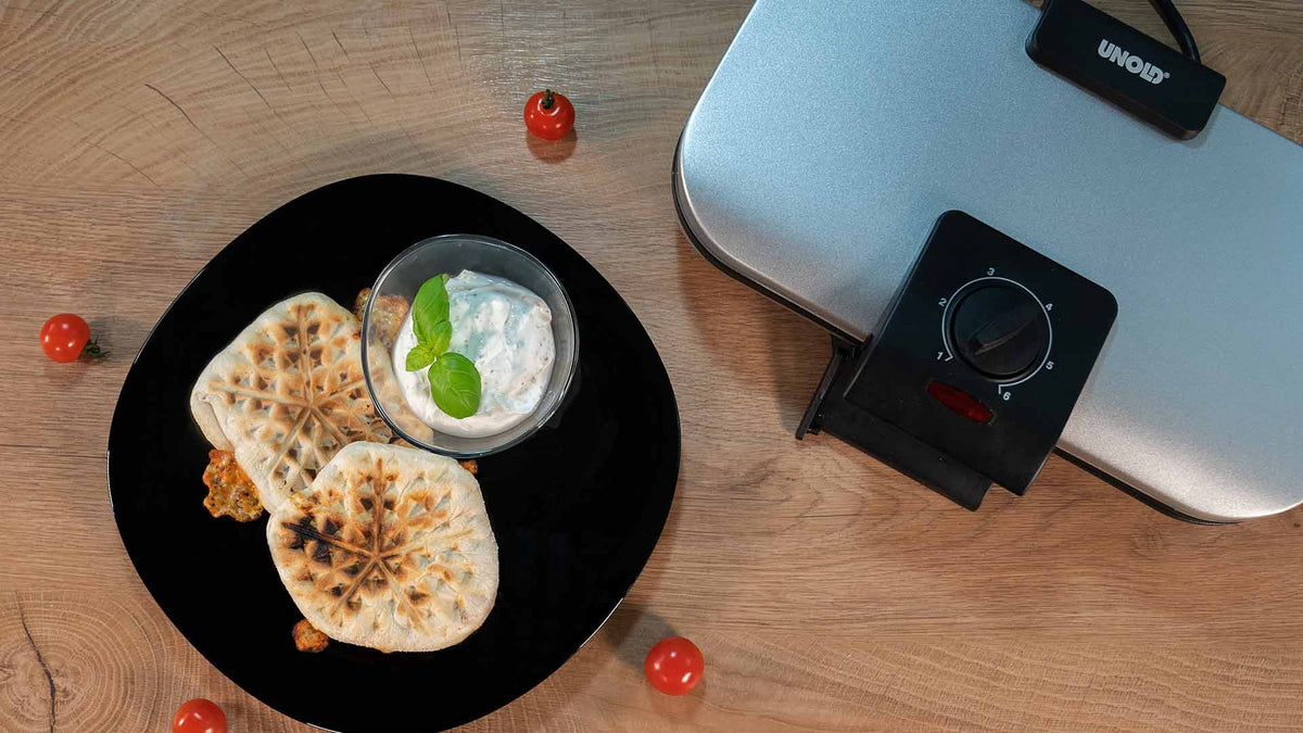 Ein schwarzer Teller mit zwei gegrillten Fladenbroten und einer Schüssel weißem Dip, garniert mit Basilikum, steht auf einem Holztisch neben einem silbernen Sandwichmaker von Uniold. Überall liegen Kirschtomaten verstreut.