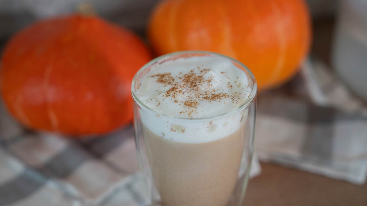 Ein Glas schaumiger Pumpkin Spice Latte mit Zimt steht auf einem karierten Tuch, im Hintergrund sind zwei orangefarbene Kürbisse zu sehen.
