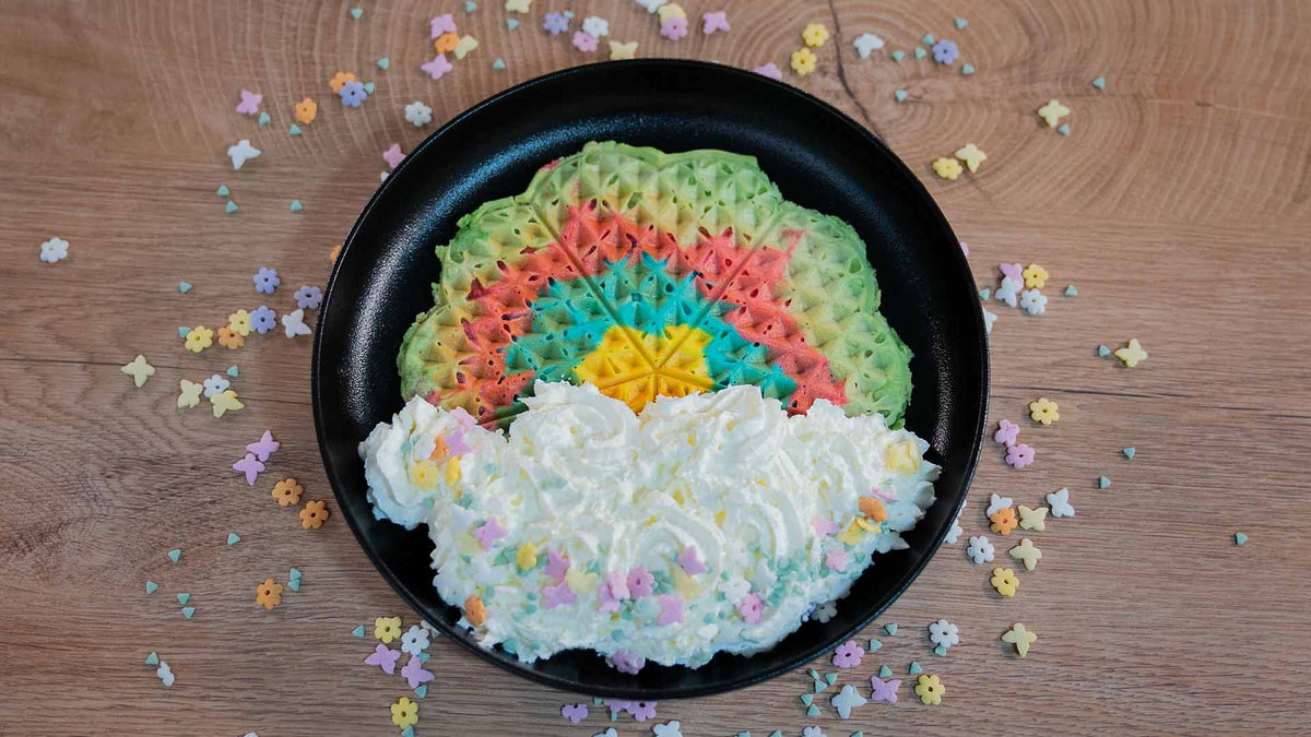 Eine regenbogenfarbene Waffel mit einem gelben Stern in der Mitte liegt auf einem schwarzen Teller. Wolkenförmige Schlagsahne und bunte Sternstreusel zieren die Waffel und umgeben den Teller auf einer Holzoberfläche.