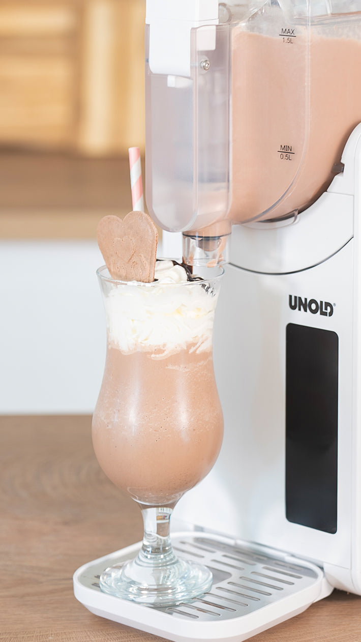 Ein Glas mit einem Schokoladenmilchshake mit Schlagsahne, einem herzförmigen Keks und einem gestreiften Strohhalm steht unter einer Milchshakemaschine auf einem Holztisch.