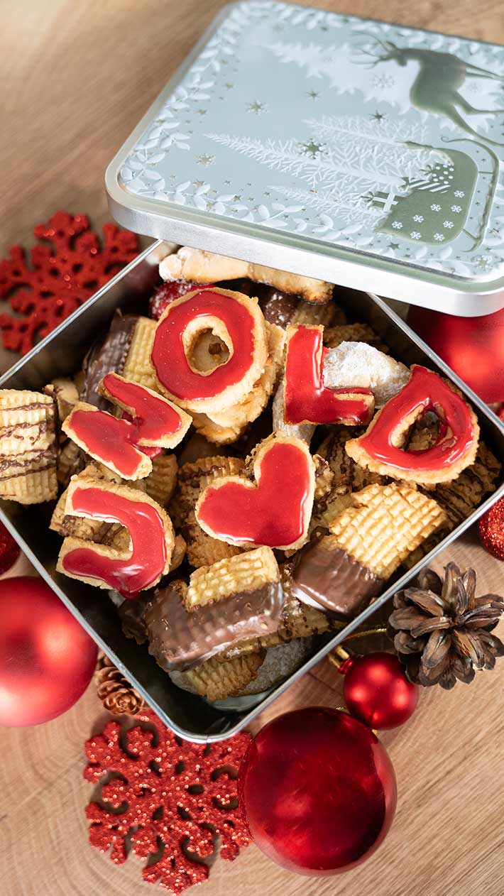 Eine offene Blechdose mit verschiedenen Keksen, einige davon mit roten Zuckergussbuchstaben, die „HOLID“ buchstabieren, und einer Herzform. Umgeben von roten Ornamenten, Tannenzapfen und festlichen Dekorationen auf einer Holzoberfläche.