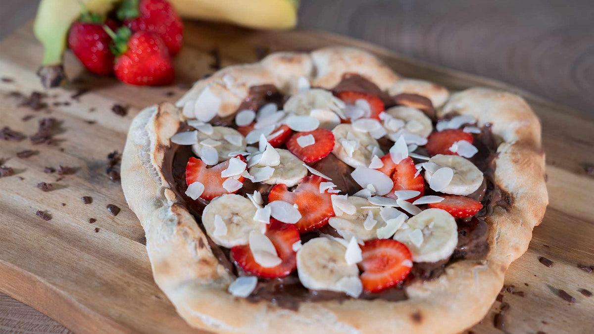 Eine rustikale Dessertpizza mit Schokoladenaufstrich, Bananenscheiben, Erdbeeren und Mandelblättchen liegt auf einem Holzbrett. Im Hintergrund sind frische Erdbeeren und Bananen zu sehen.