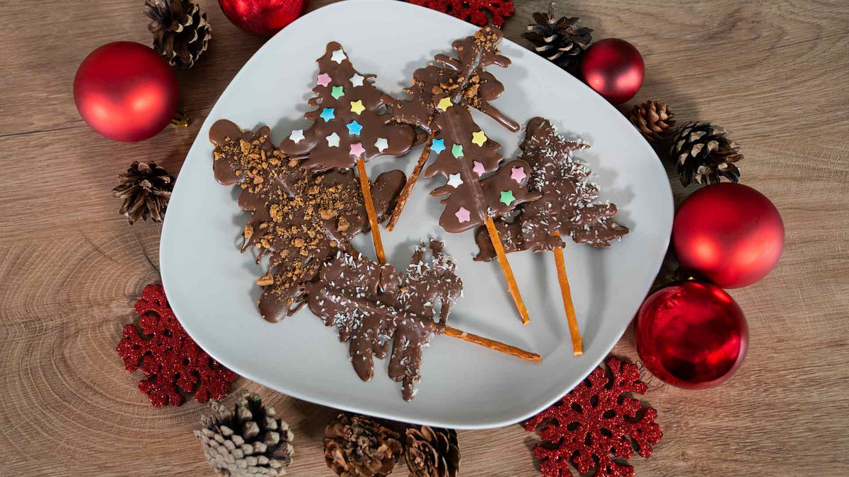 Auf einem weißen Teller liegen Schokoladen-Weihnachtsbaumleckereien aus Salzstangen, verziert mit bunten Streuseln, umgeben von roten Ornamenten, Tannenzapfen und roten Schneeflockendekorationen auf einer Holzoberfläche.