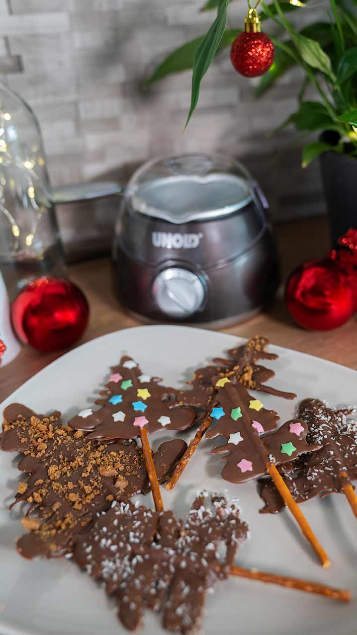 Ein Teller mit Schokoladen-Weihnachtsbaum-Leckereien auf Brezelstangen, dekoriert mit bunten Streuseln, Kokosnuss und Zimt, steht auf einem Tisch mit roten Ornamenten und einem Schokoladenschmelzer im Hintergrund.
