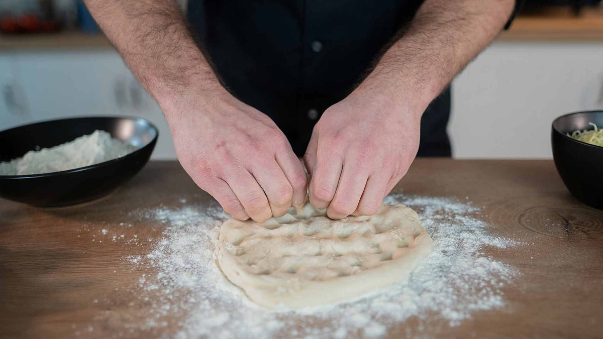 Eine Person presst ihre Fingerknöchel auf einer bemehlten Holzoberfläche zu einer runden Kugel Pizzateig. Daneben stehen Schüsseln mit Mehl und Zutaten.