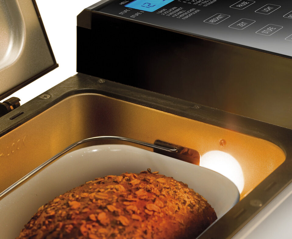 Eine Nahaufnahme des Unold Backmeister® Top Edition Brotbackautomaten mit geöffnetem Deckel zeigt ein im Inneren backendes Körnerbrot. Das digitale Bedienfeld und die Tasten befinden sich auf der rechten Seite des Geräts.