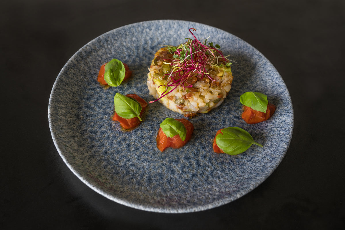 Das Unold Raclette Rezeptbuch Deluxe präsentiert sich auf einem blaugrauen Teller als runde Risottoportion mit Microgreens, umgeben von vier Klecksen Tomatensoße, jeweils garniert mit Basilikum, vor dunklem Hintergrund.